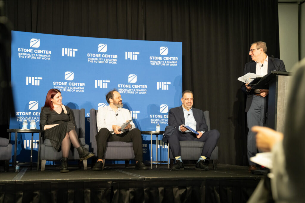Three panelists sit in armchairs onstage, smiling. A fourth speaker stands at the podium, turning back toward the other three panelists.