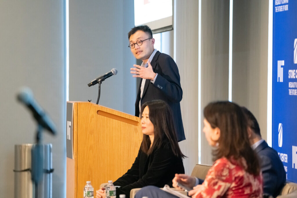 A speaker stands at a wooden MIT podium, gesturing while presenting. Two panelists are seated in the foreground, partially out of focus.